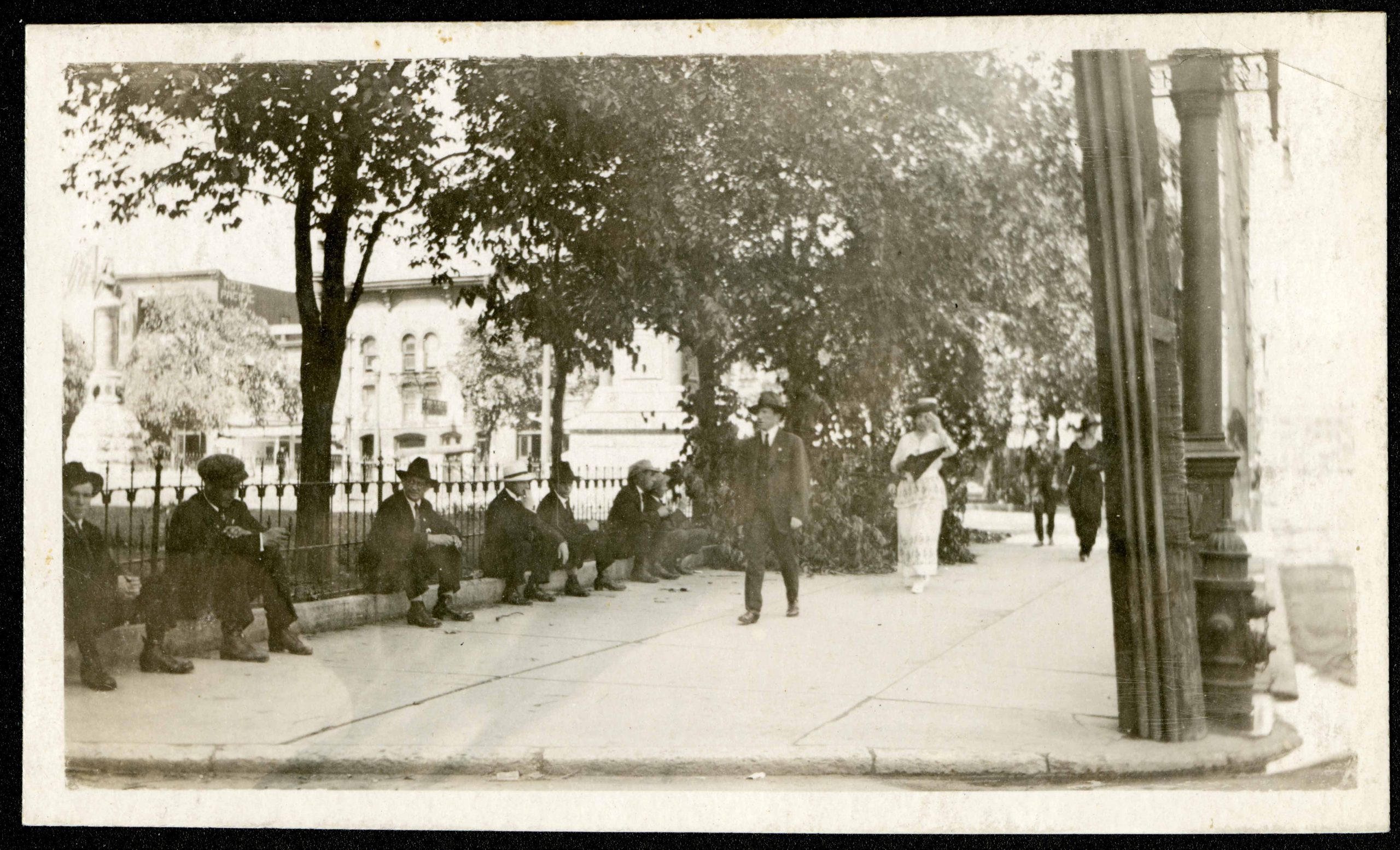 1924-Photograph of men sitting on the sidewalk in front of City Hall while others walk passed them